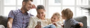 A dad, mom, little girl, and little boy sitting on the couch smiling and looking at a phone