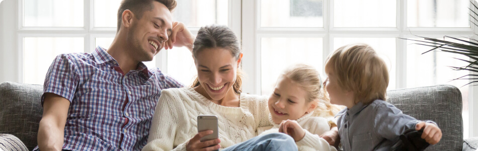 A dad, mom, little girl, and little boy sitting on the couch smiling and looking at a phone